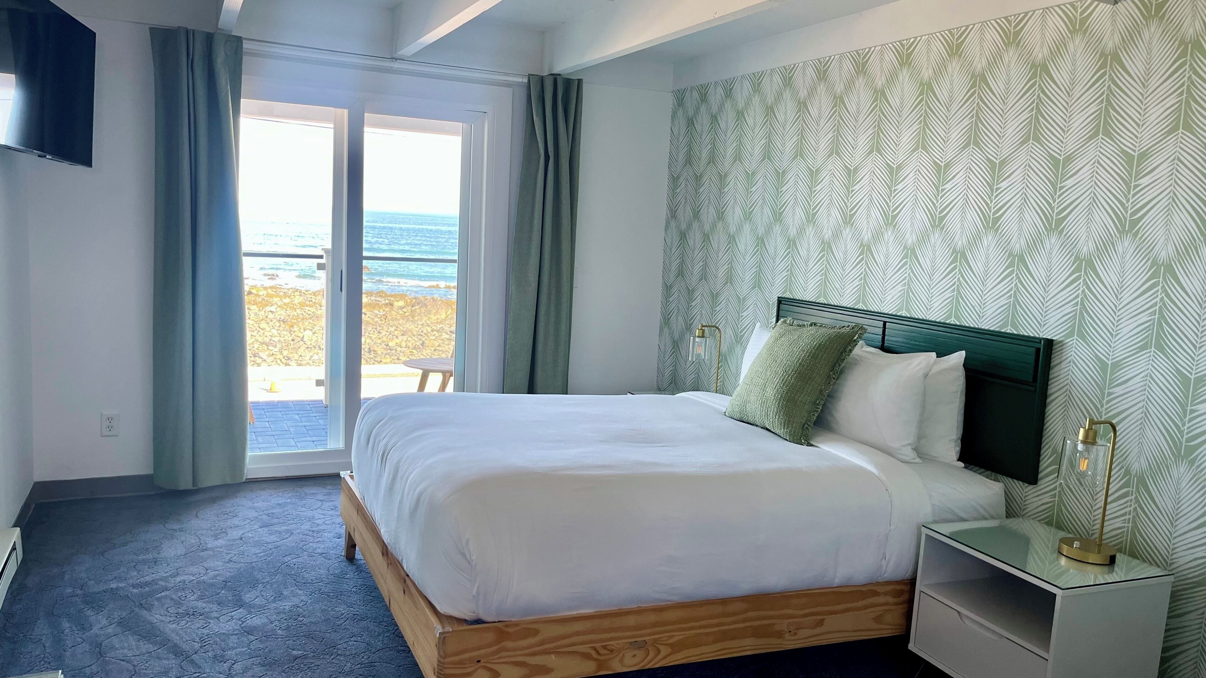 A bedroom with a bed, a nightstand, a lamp, a window with a view of the beach, and a patterned wall.