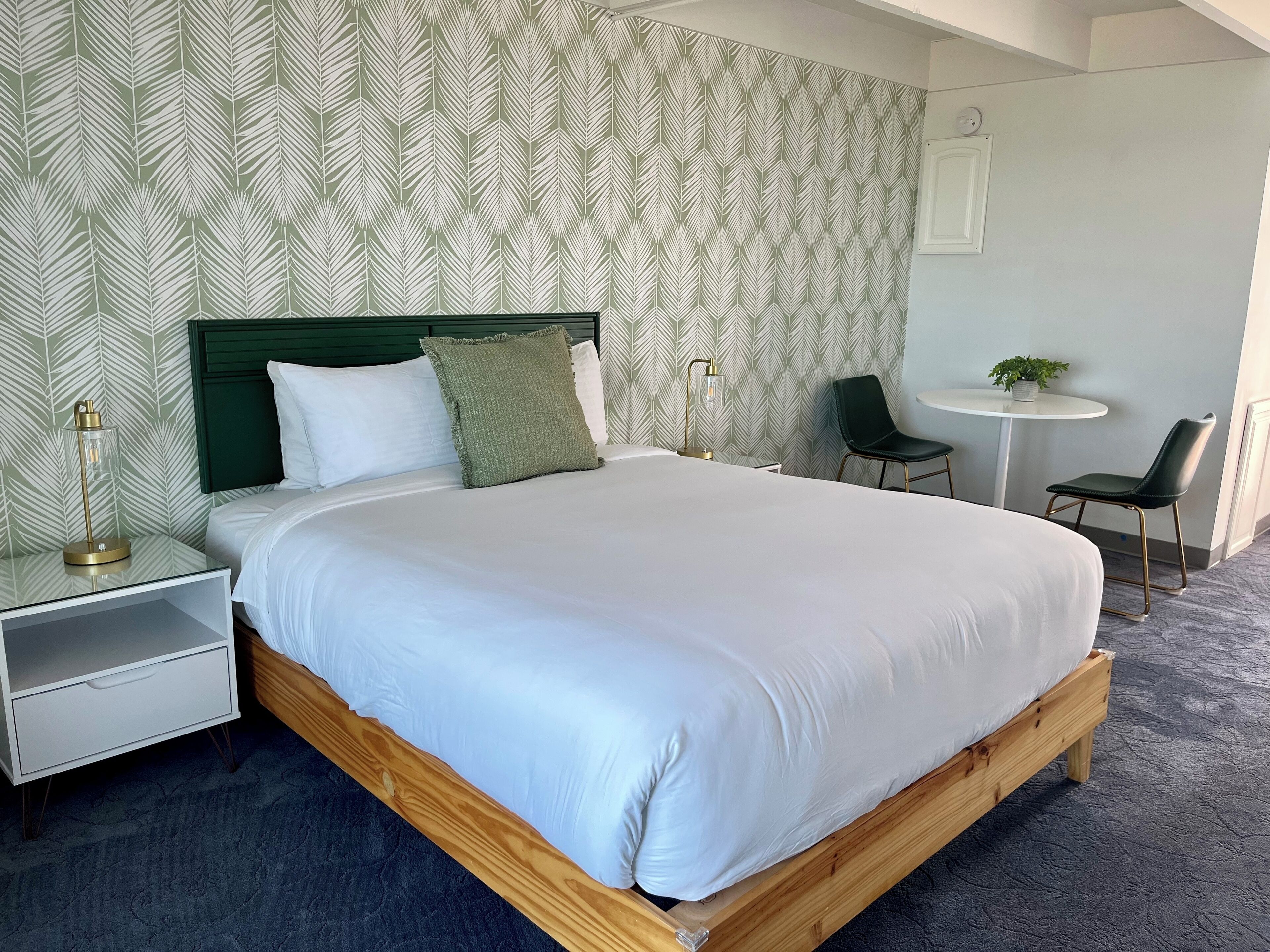 A neatly arranged bedroom with a wooden bed, white bedding, a green pillow, a bedside lamp, a small white nightstand, and a wall with a leaf-pattern wallpaper.