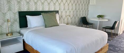 A neatly arranged bedroom with a wooden bed, white bedding, a green pillow, a bedside lamp, a small white nightstand, and a wall with a leaf-pattern wallpaper.
