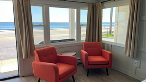 A room with two orange chairs, a small round table, and a view of the beach through large windows. - The Grandview Hotel (York Beach)