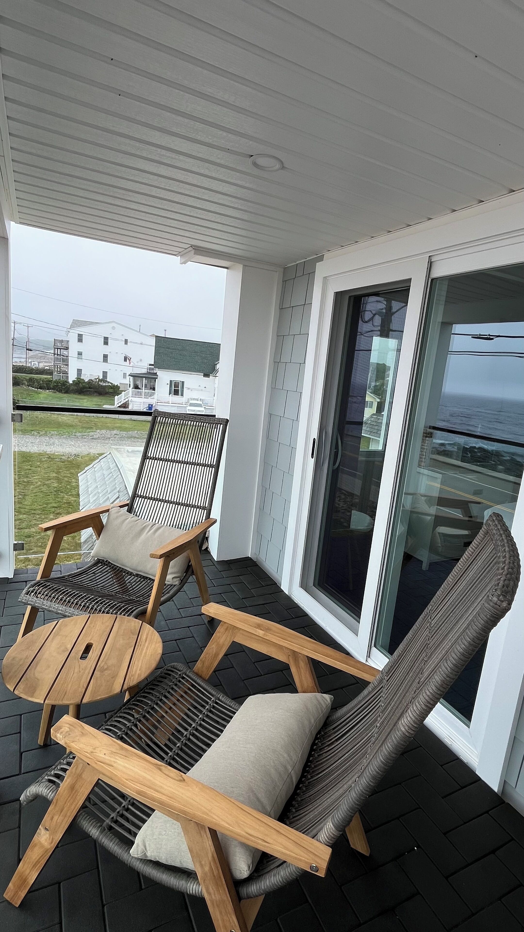 A covered outdoor seating area with wicker chairs and a round wooden table, overlooking a residential area and the sea.