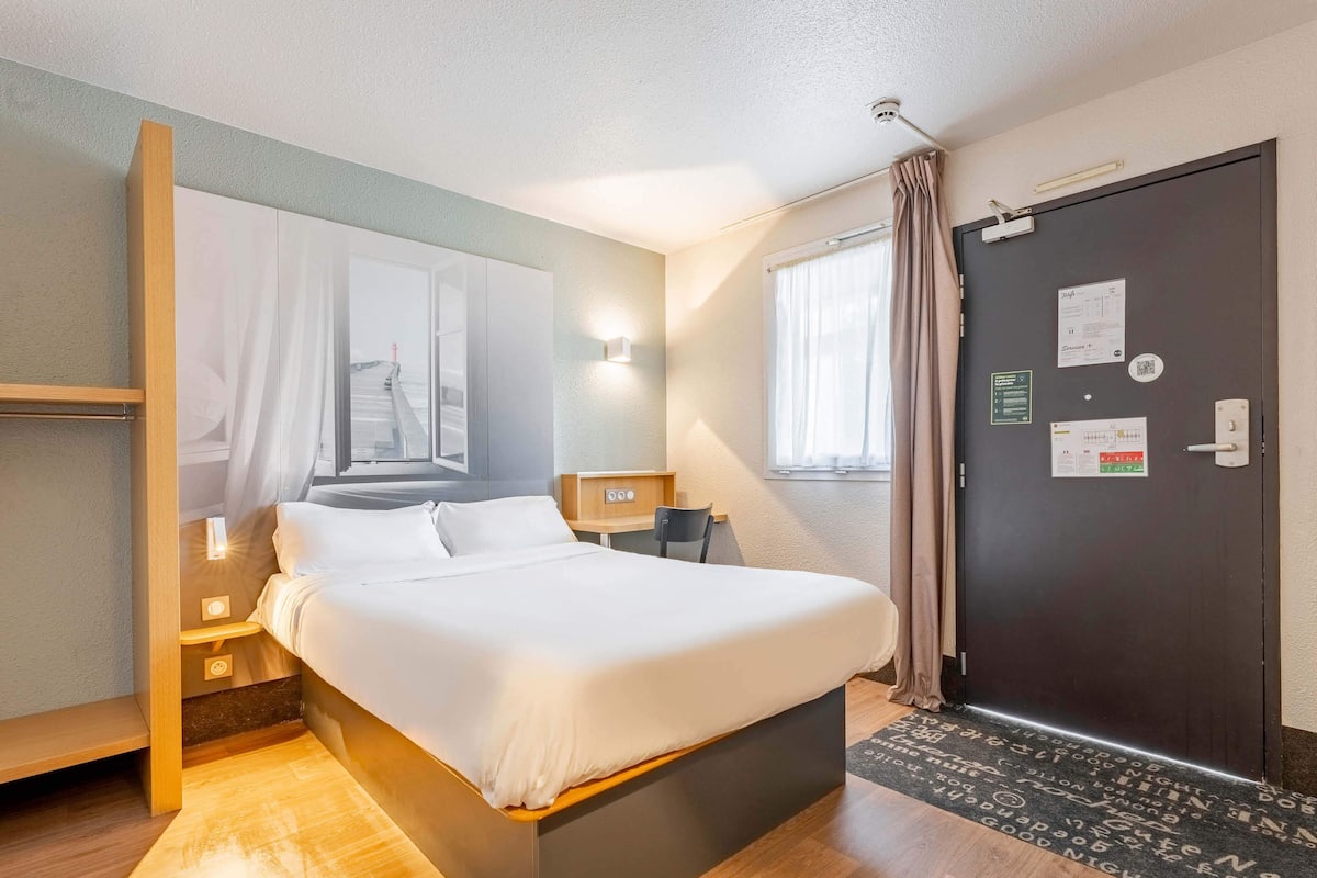 Ein Hotelzimmer mit Bett, Nachttisch, Lampe und einer Tür mit einem Korkbrett.