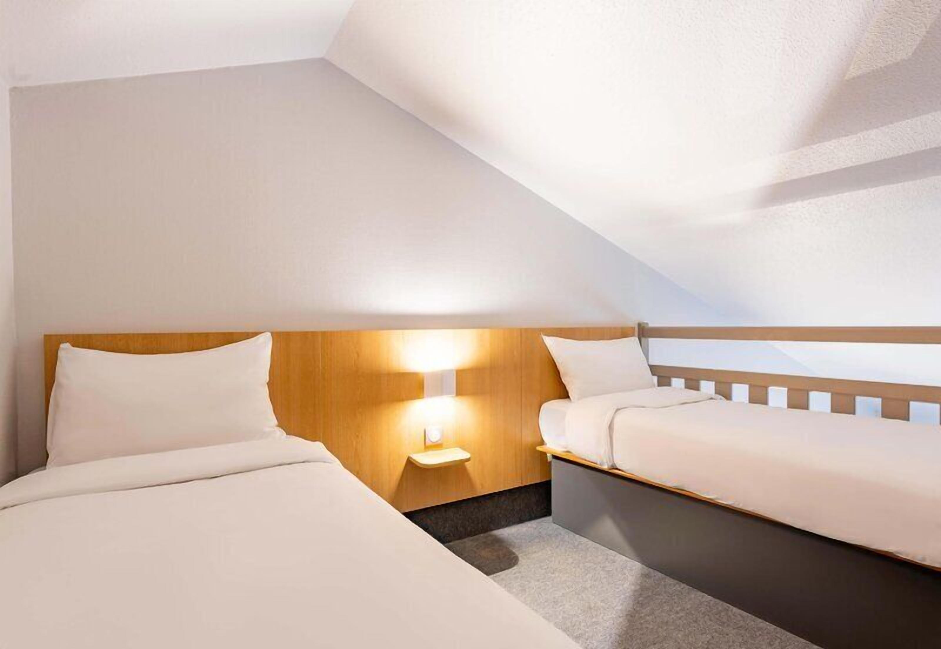 Une chambre d’hôtel avec deux lits, une tête de lit en bois, une table de chevet avec une lampe et un plafond en pente.