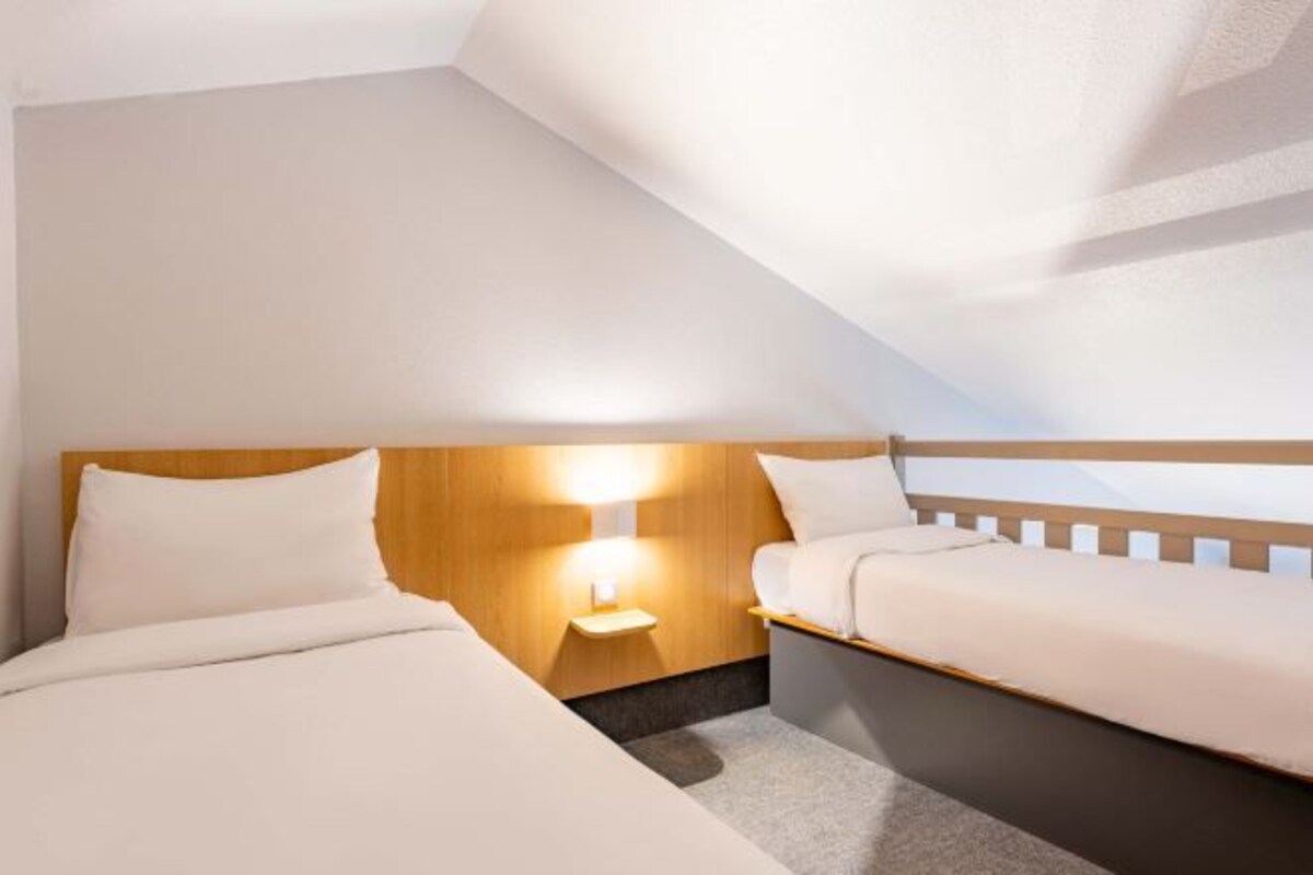 A hotel room with a sloped ceiling, two beds with white bedding, a wooden headboard, and a bedside table with a lamp.