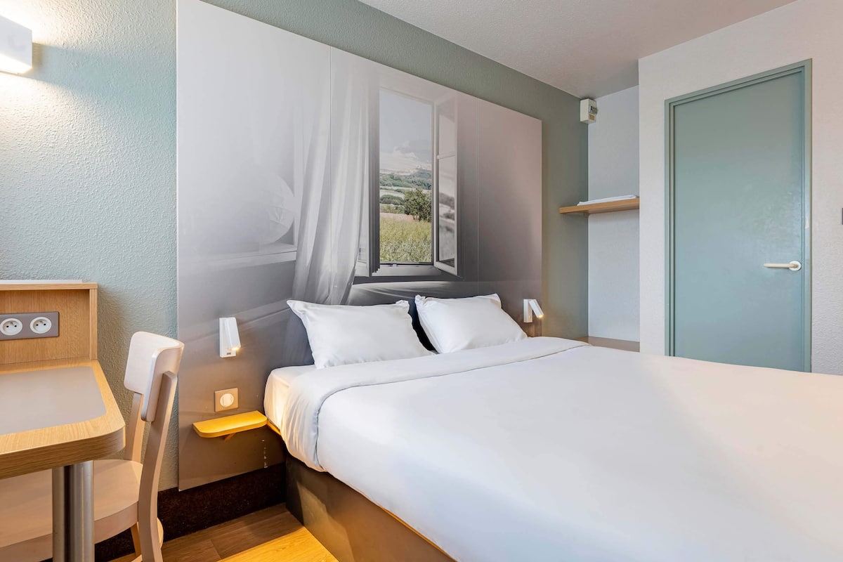 Ein Hotelzimmer mit Bett, Schreibtisch, Stuhl, einem Fenster mit Vorhängen und einer Tür.