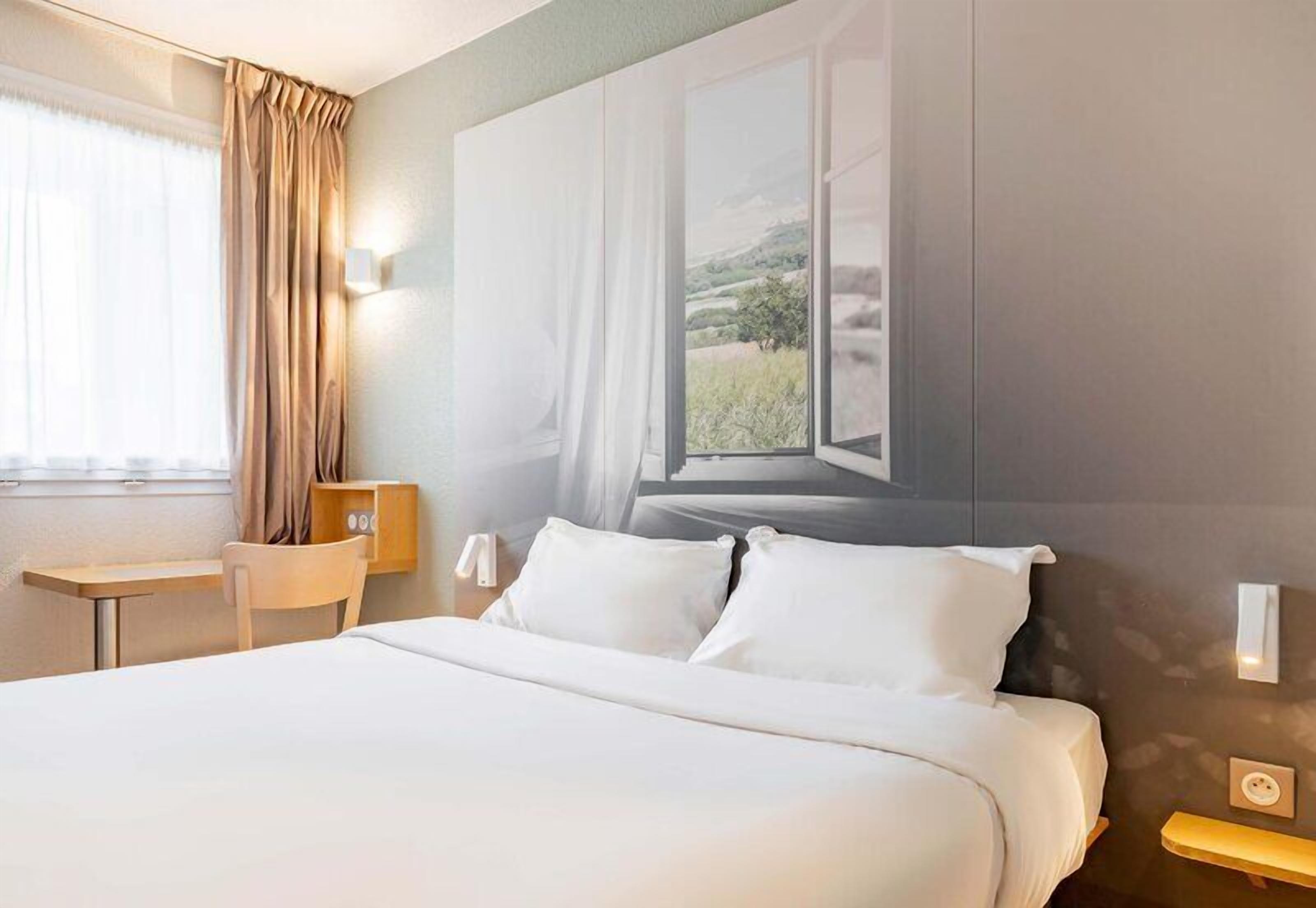A hotel room with a bed, a desk, a chair, a window with curtains, and a wall-mounted lamp.