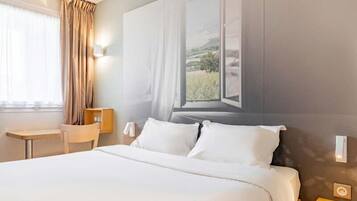 A hotel room with a bed, a desk, a chair, a window with curtains, and a wall-mounted lamp.
