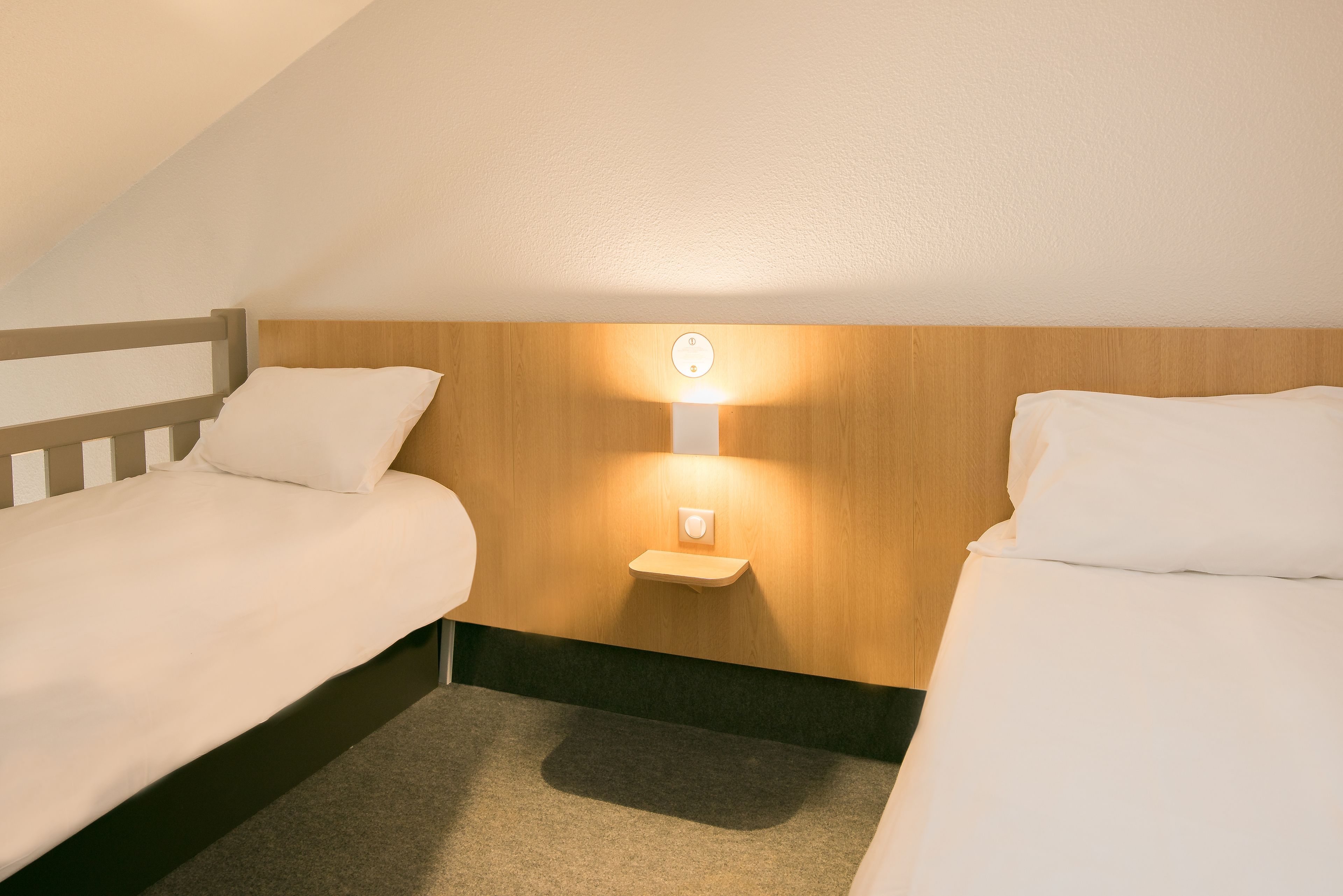 Une chambre d’hôtel avec deux lits simples, une tête de lit en bois, une table de chevet avec une lampe et un sol recouvert de moquette.