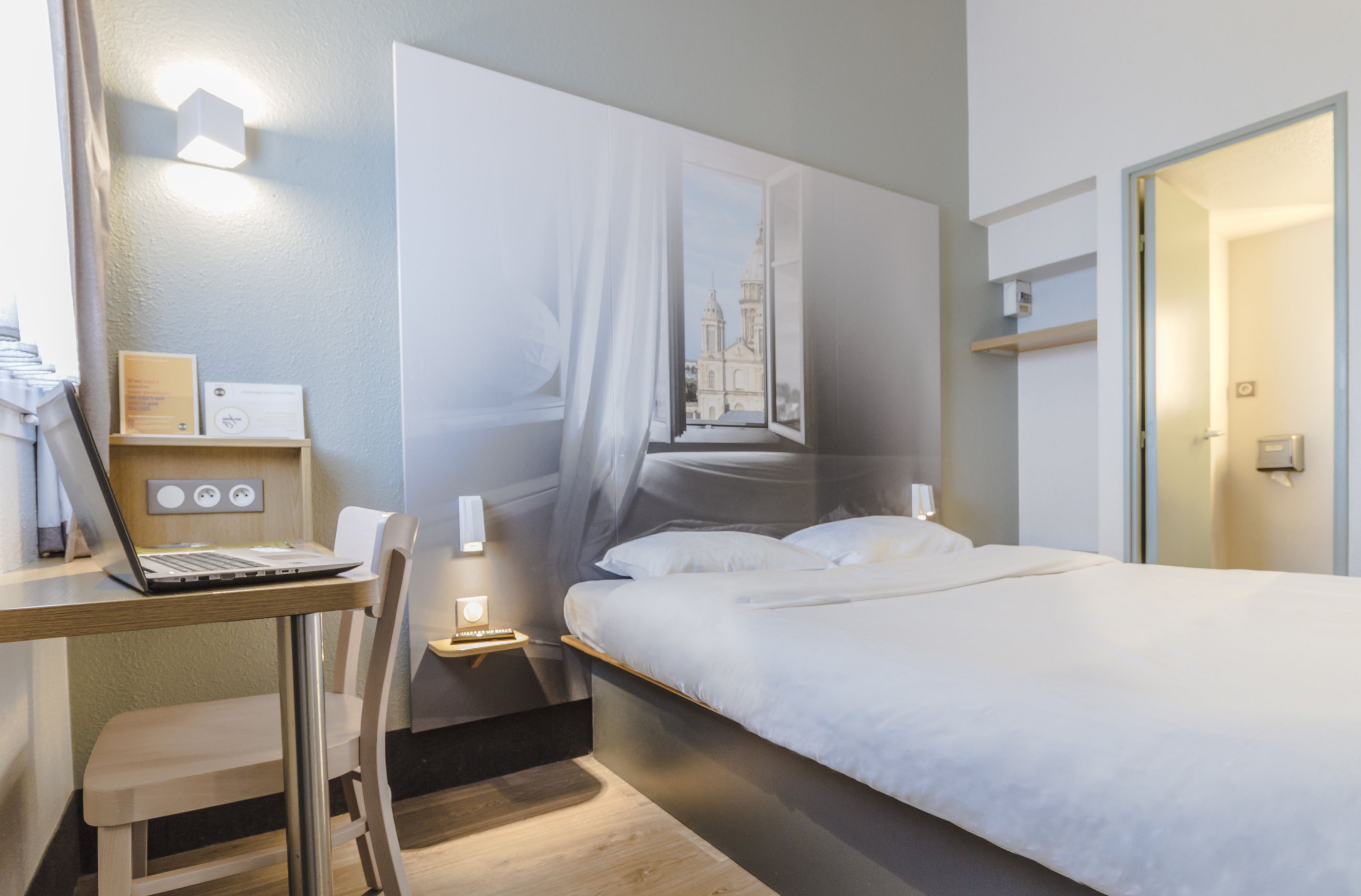 Habitación de hotel moderna con una cama, un escritorio con una computadora portátil y vistas a un edificio a través de una ventana.