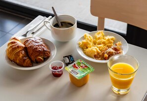 Una mesa de desayuno con cruasanes, huevos revueltos, un vaso de zumo de naranja, una taza de café y un tarro pequeño de mermelada.