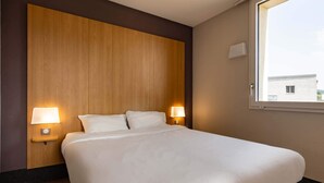 Habitación de hotel con una cama grande, dos lámparas de noche, un cabecero de madera y una ventana con vistas a edificios.