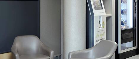 Un interior moderno con dos sillones de cuero gris, una mesita redonda, un quiosco de autoservicio y un refrigerador lleno de bebidas.