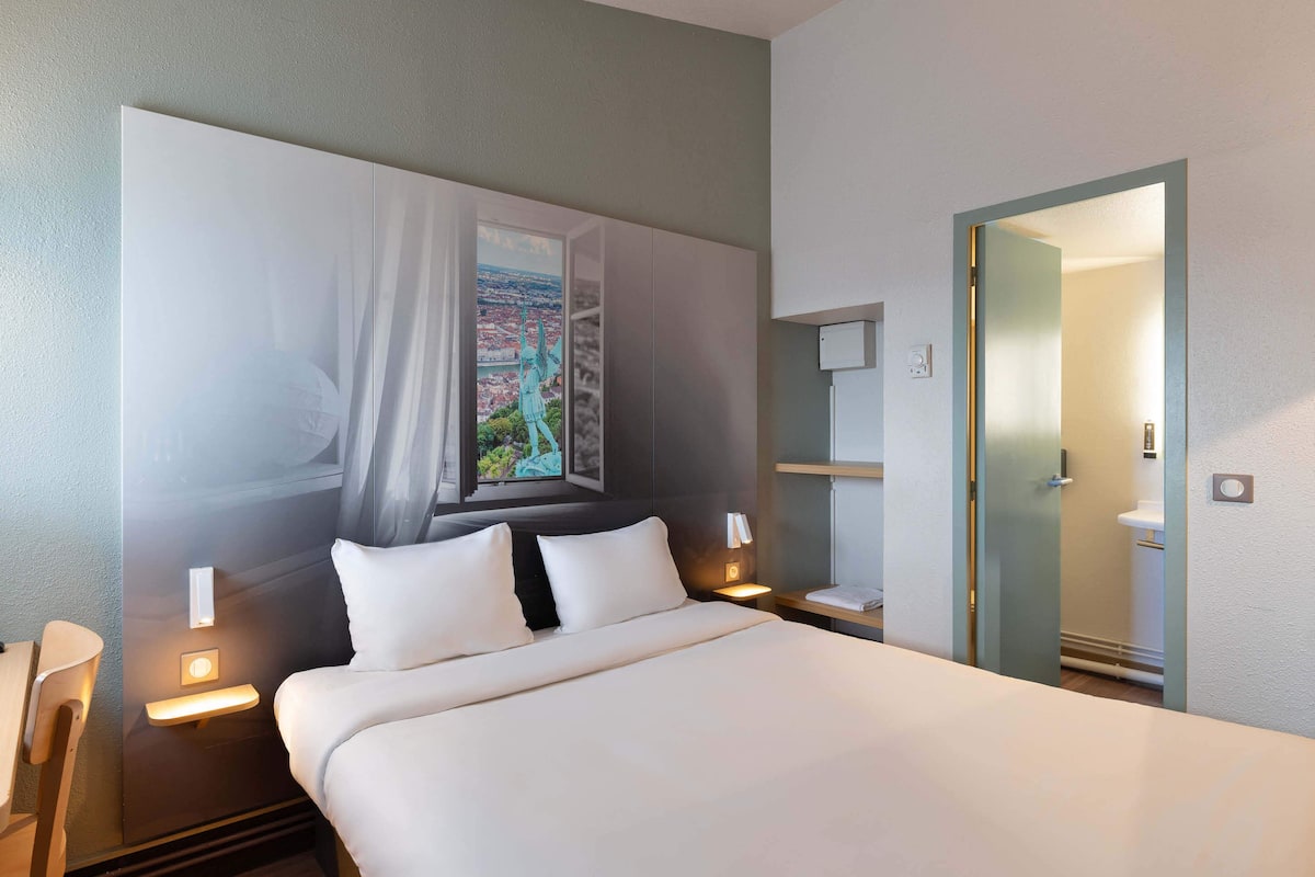 A hotel room with a large bed, a nightstand with a lamp, a wall-mounted shelf, and a door leading to a bathroom.