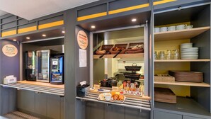 A breakfast counter with a variety of food items and utensils, a coffee machine, and a display of plates and bowls. - B&B HOTEL Lyon Saint-Priest (Saint-Priest)