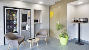 A modern waiting area with a vending machine, two chairs, a small table, a potted plant, and a microwave on a shelf. - B&B HOTEL Lyon Saint-Priest (Saint-Priest)