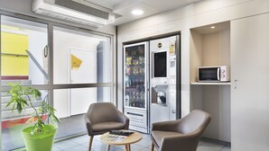 A modern indoor area with two brown chairs, a green potted plant, a vending machine, and a microwave. - B&B HOTEL Lyon Saint-Priest (Saint-Priest)