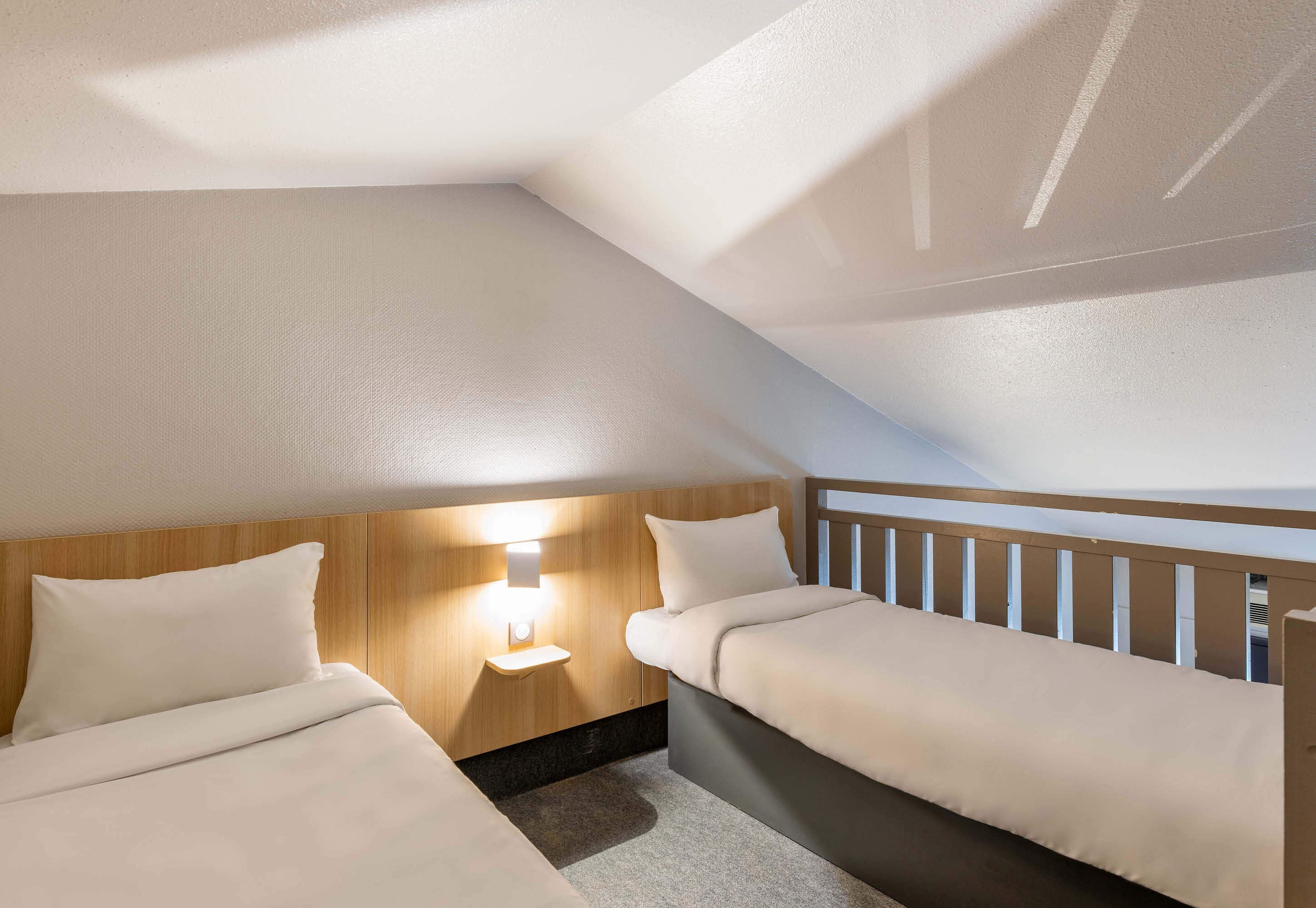 A room with two beds, a wooden headboard, a nightstand with a lamp, and a view of the sky.