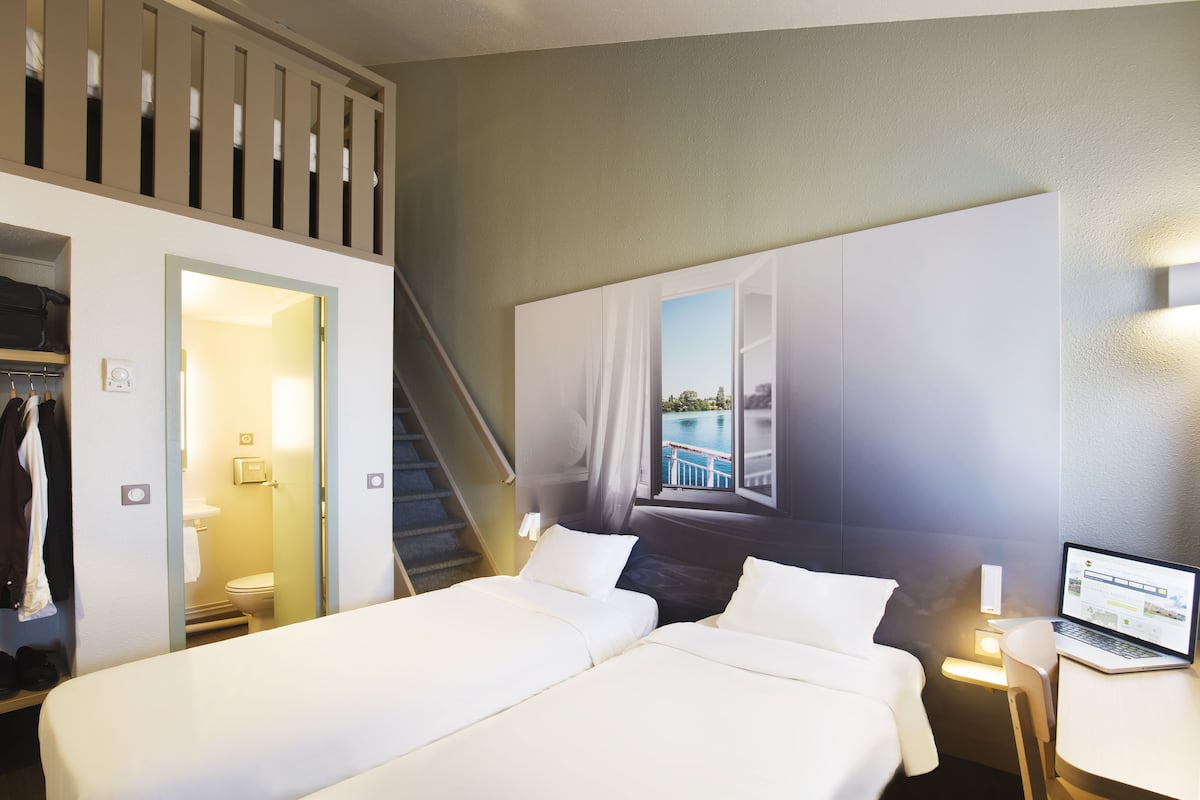 A hotel room with two beds, a laptop on a bedside table, a staircase, and a view of a pool through a window.