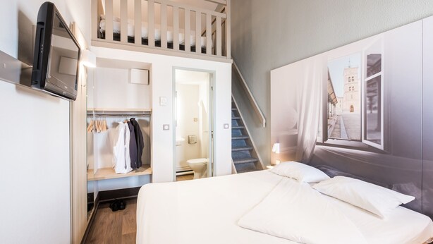 A hotel room with a bed, a TV, a closet, a staircase, and a view of a building through a window. - B&B HOTEL Lyon Saint-Priest (Saint-Priest)