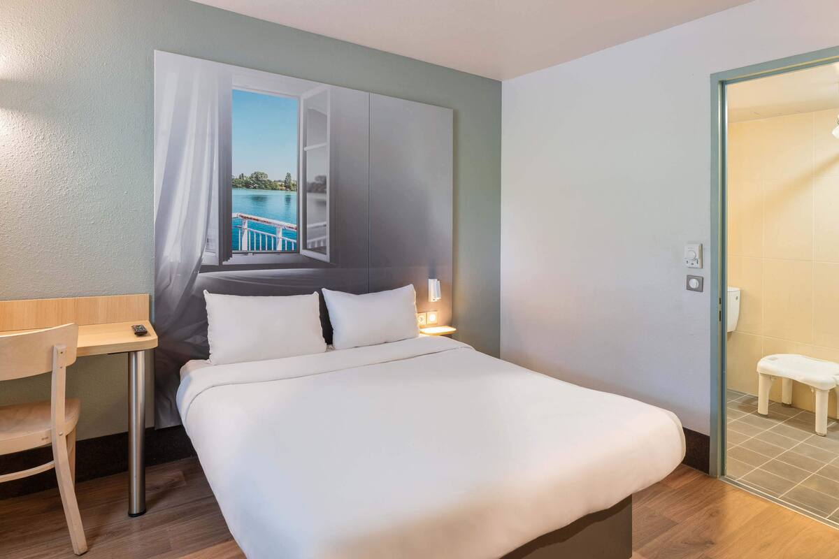 A hotel room with a bed, a desk, a chair, a nightstand, and a bathroom visible through an open door.
