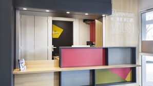 A reception desk with a 'Bienvenue Chez Vous' sign, a counter, and a view of the interior. - B&B HOTEL Lyon Saint-Priest (Saint-Priest)