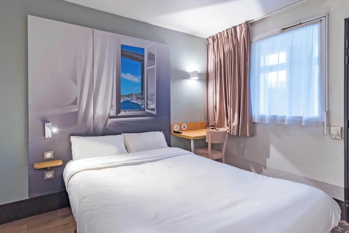 Une chambre d’hôtel comprenant un lit, un bureau, une chaise, une fenêtre avec des rideaux et un luminaire fixé au mur.