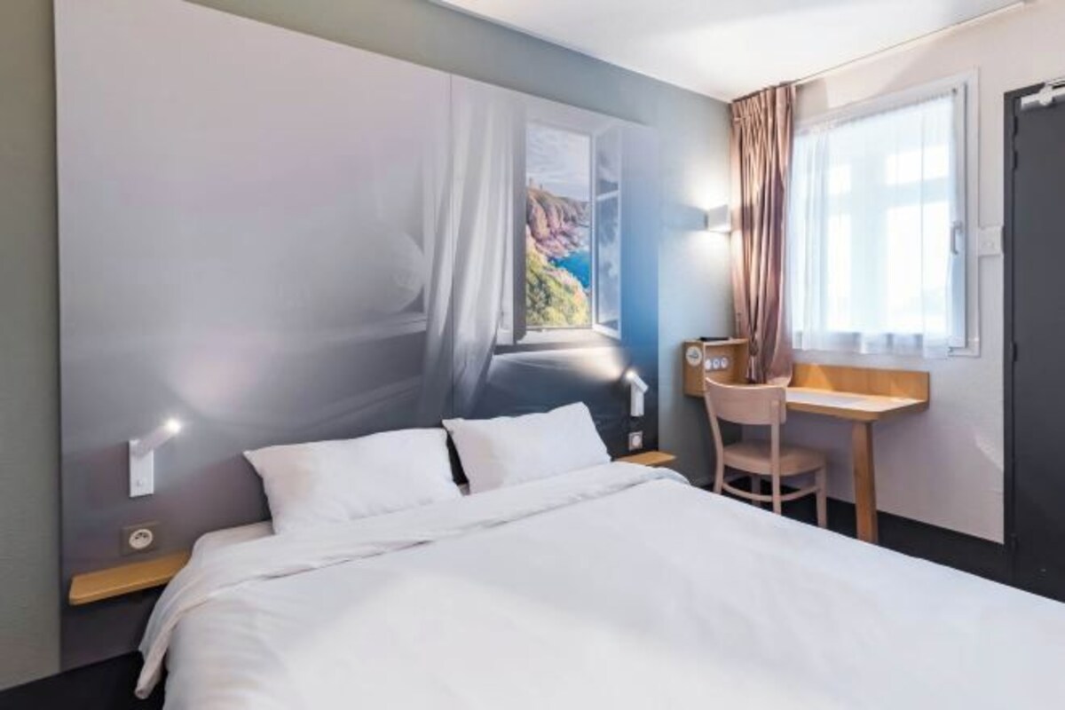 A hotel room with a bed, a desk, and a chair. There is a window with curtains, and a wall mural depicting a scenic view.