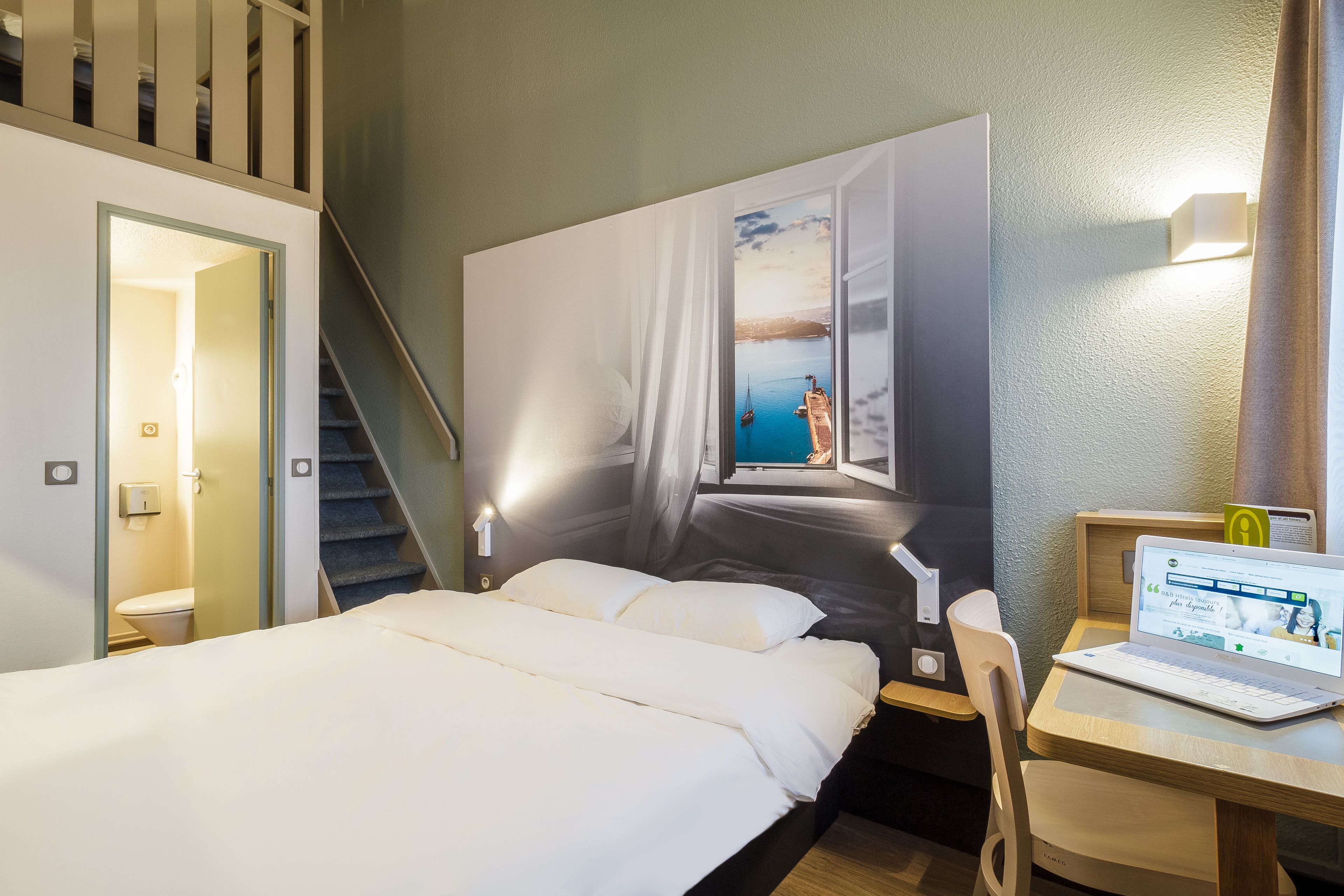 Habitación de hotel con cama, escritorio con una computadora portátil, ventana grande con vista al mar y una escalera.