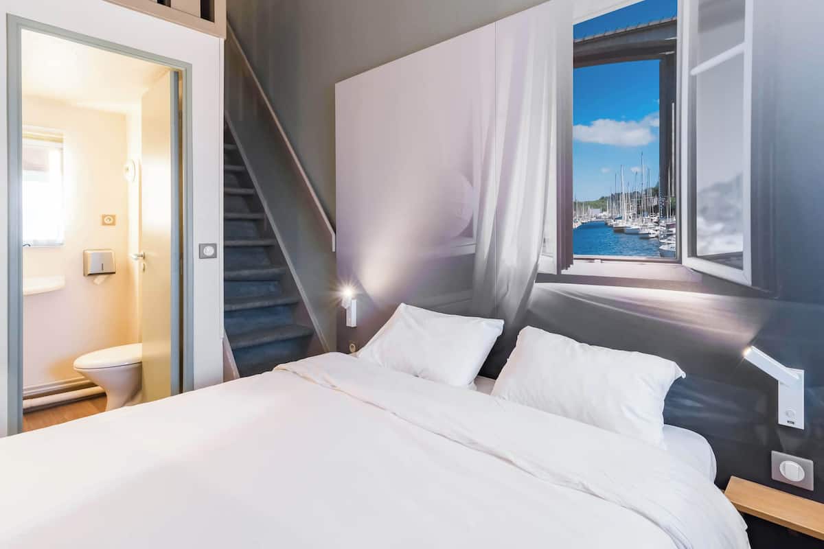 Une chambre à coucher avec un lit, une fenêtre donnant sur un port de plaisance, un escalier et une porte menant à une salle de bain.