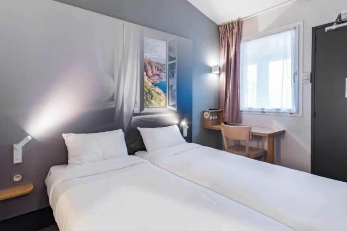 A hotel room with a bed, a desk, a chair, a window with curtains, and a wall-mounted light.