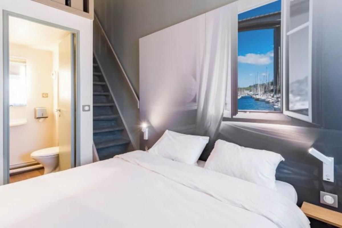 A modern hotel room with a bed, a window with a view of boats, and a staircase.