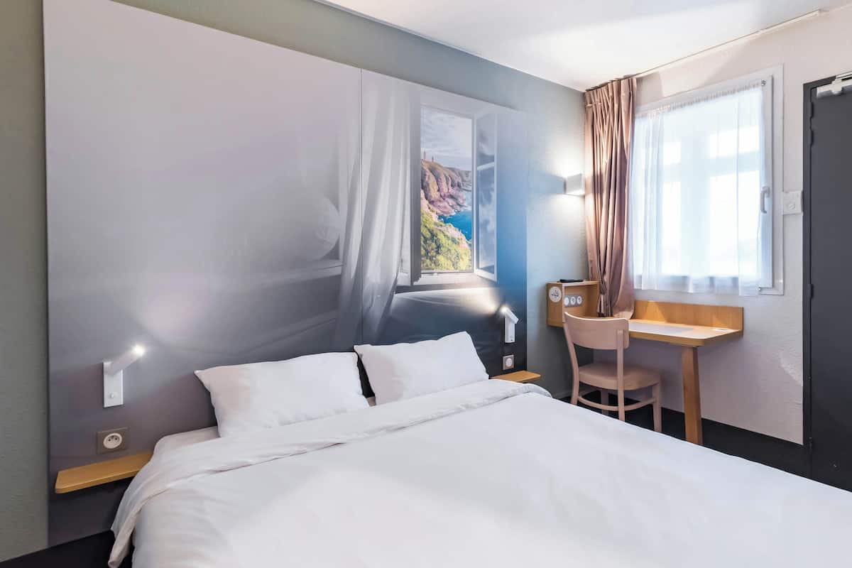 Une chambre d’hôtel avec un lit, un bureau, une chaise et une vue sur un paysage côtier, visible par une fenêtre ouverte.