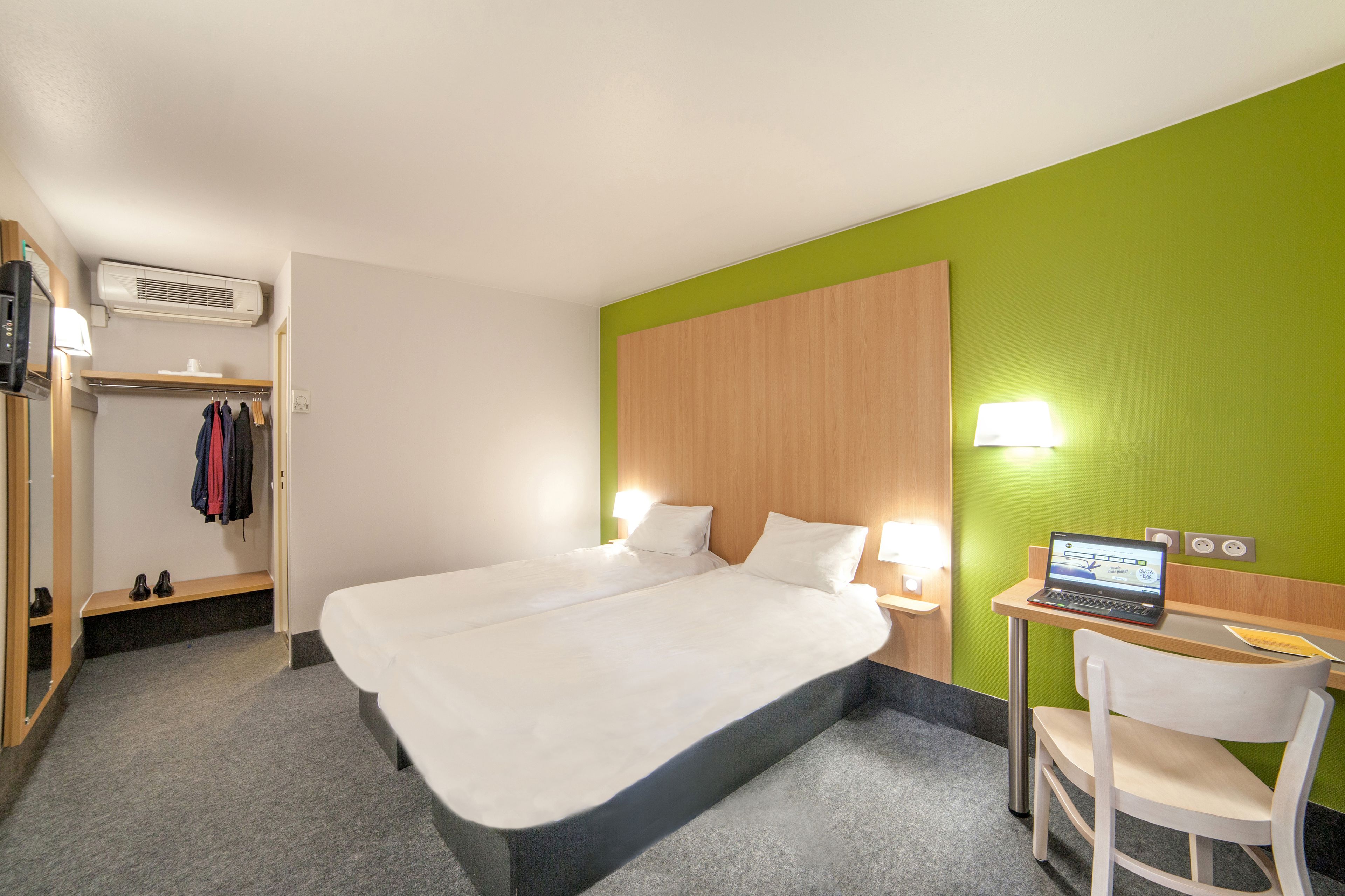 A hotel room with a large bed, a desk with a laptop, a chair, a wardrobe, and a mirror.