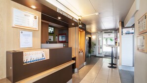 Ett välkomstområde med en reception i trä, en skylt med texten ”Välkommen till B.B.” och ett inramat certifikat på väggen.