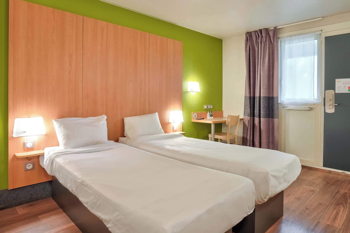 Ein Hotelzimmer mit zwei Betten, einem Holz-Kopfteil, einer grünen Akzentwand, einem kleinen Tisch mit Stuhl und einem fenster mit Vorhängen.