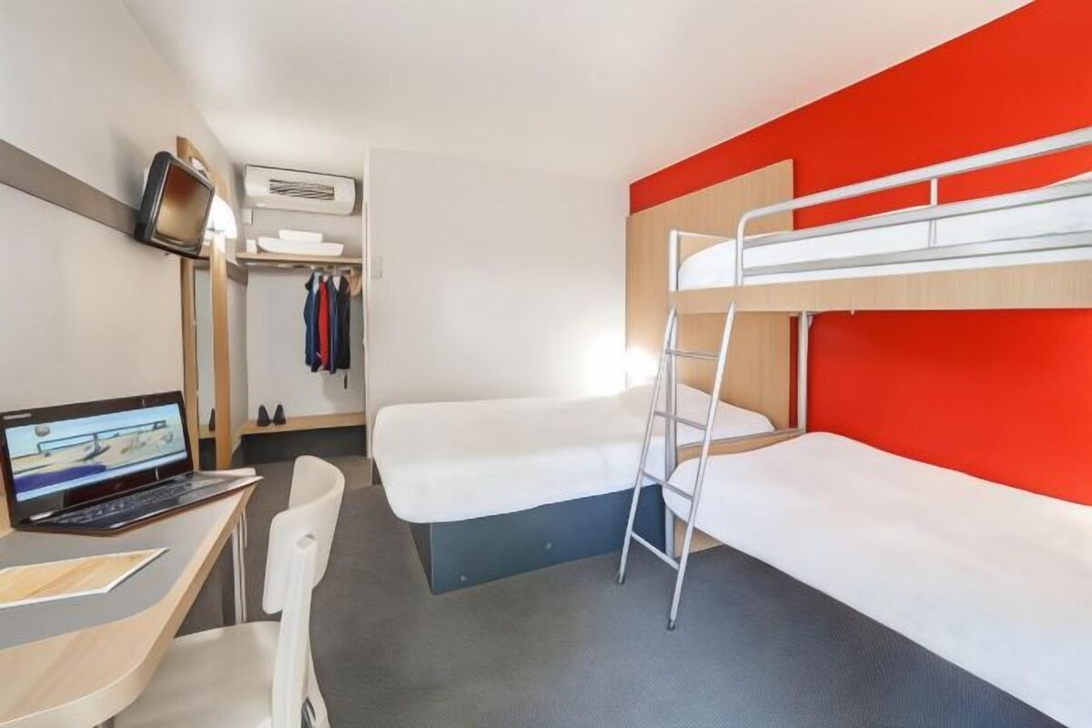 A compact hotel room with a bunk bed, a desk with a laptop, a chair, and a TV mounted on the wall.