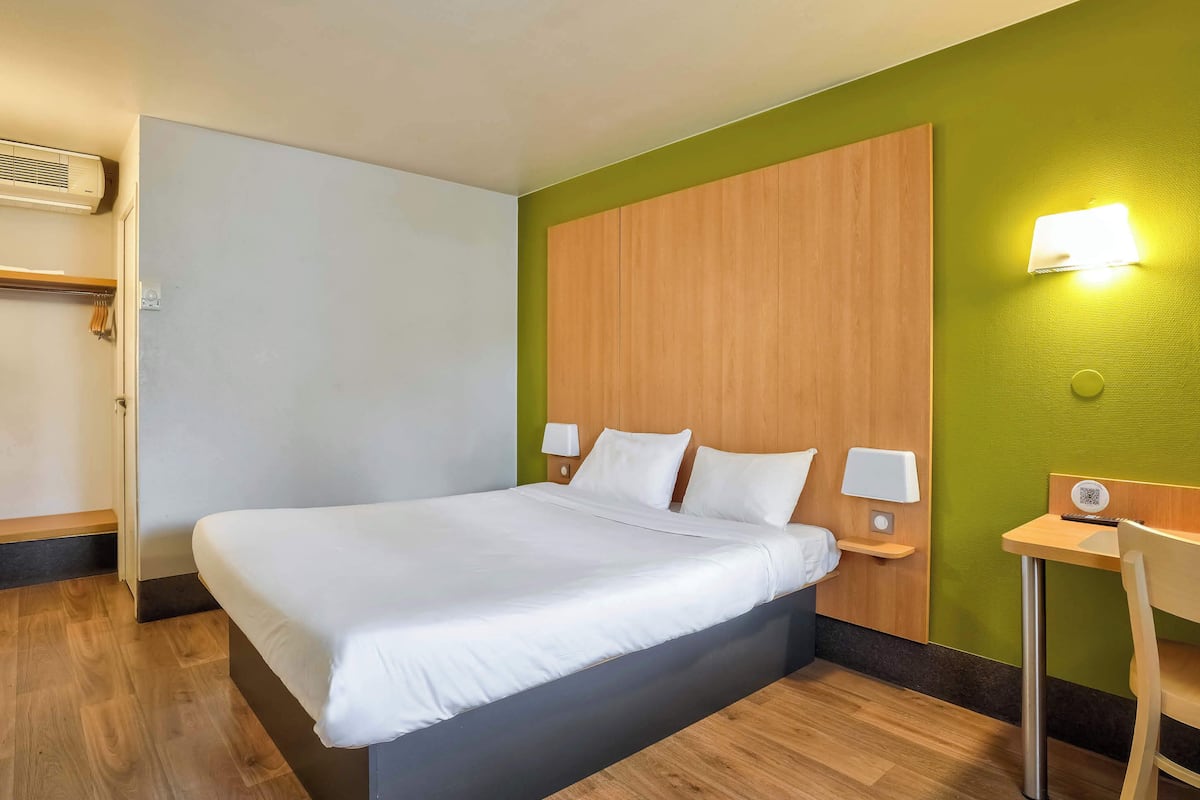 Ein Hotelzimmer mit einem großen Bett, einem Holz-Kopfteil, einem Nachttisch mit Lampe sowie einem kleinen Schreibtisch und Stuhl.
