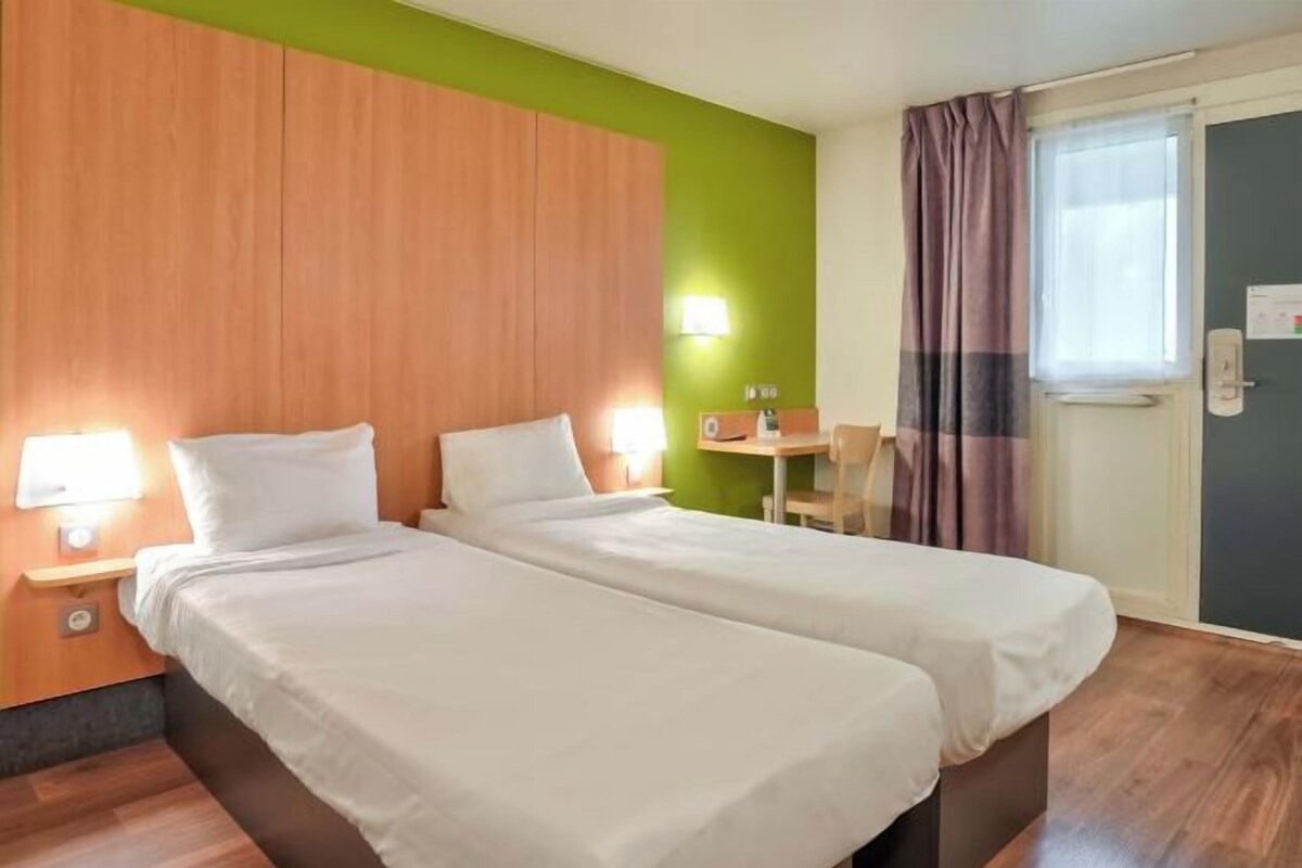 Habitación de hotel con dos camas individuales, un cabecero de madera, una pared de acento verde, una mesita con una silla y una ventana con cortinas.