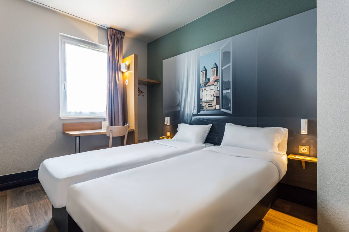 A double bed with white linens, a nightstand with a lamp, a window with curtains, and a wall-mounted painting of a cityscape.
