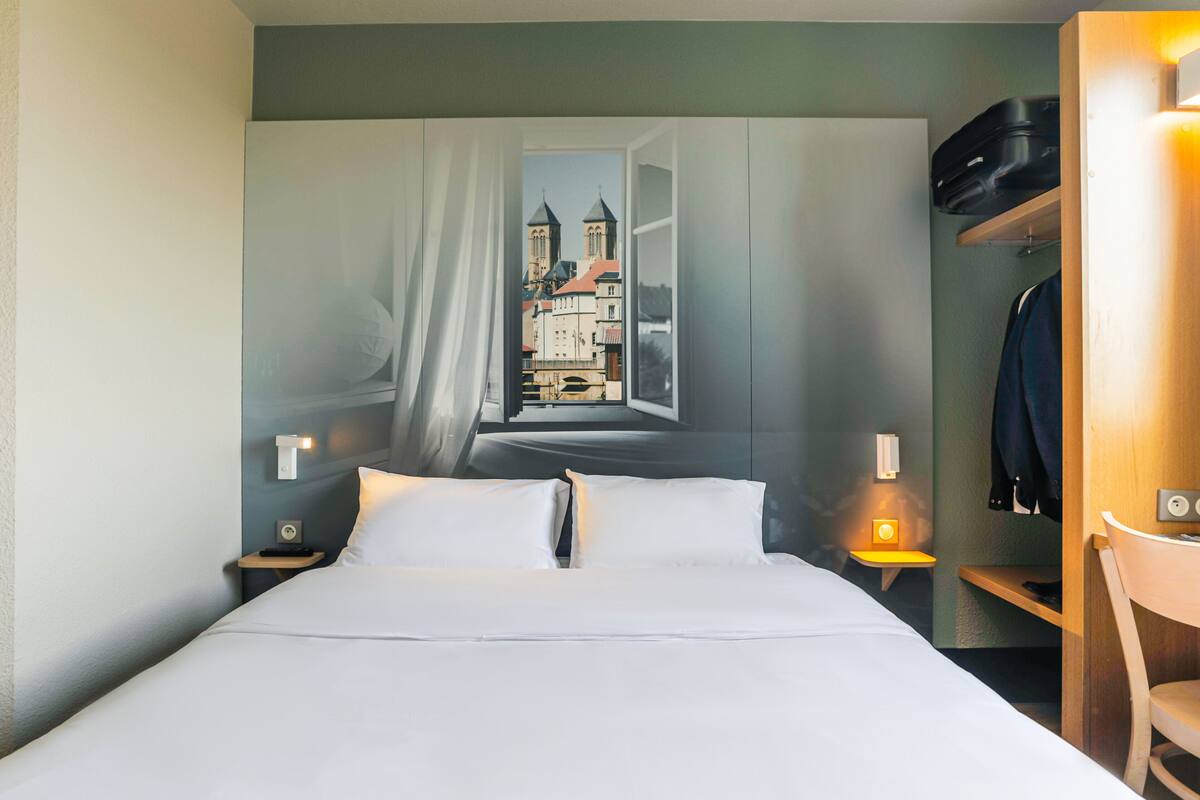 A hotel room with a bed, a nightstand, a chair, and a coat rack.