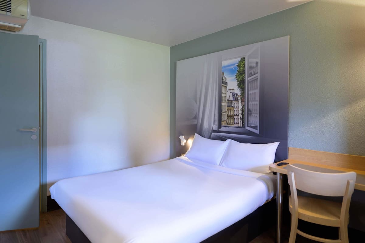 A hotel room with a bed, a desk, a chair, and a painting of a cityscape on the wall.