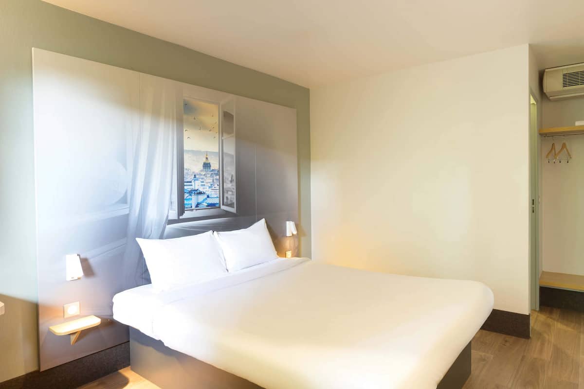 A hotel room with a large bed, a nightstand, a wall-mounted air conditioner, and a window with a cityscape view.
