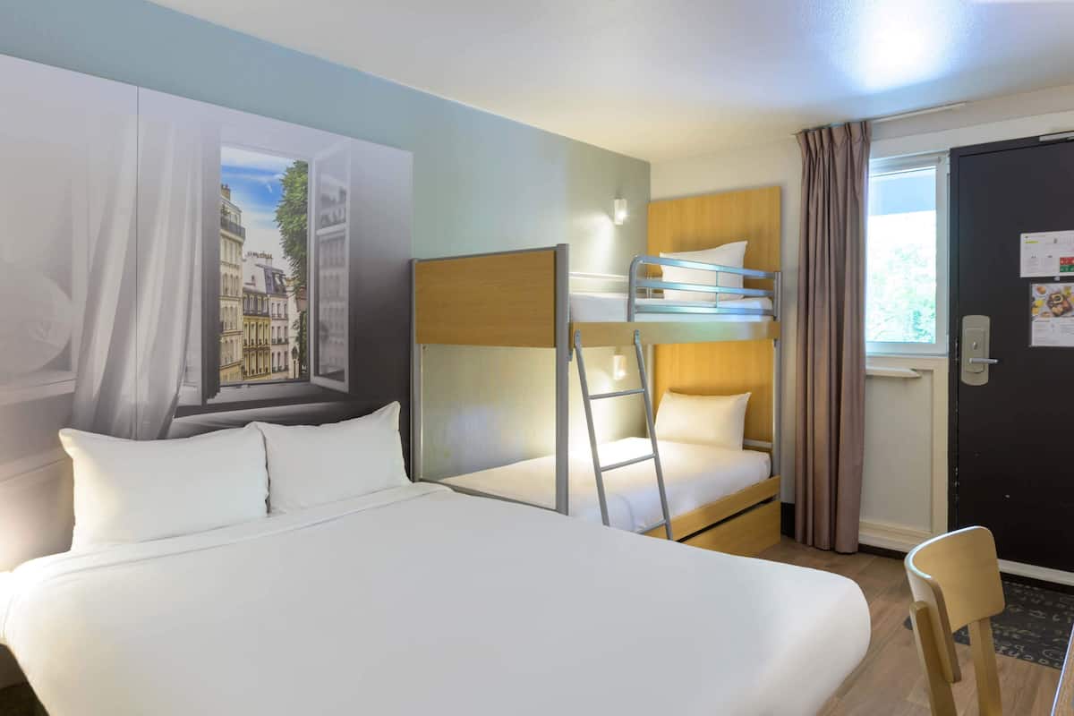 A hotel room with a large bed, a bunk bed, a desk, and a window with curtains.