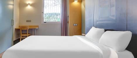 A hotel room with a bed, a desk, a chair, a window with curtains, and wall-mounted lights.