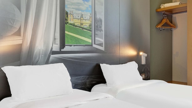 A hotel room with a bed, a window with a view of a building, a wooden hanger, and a wall-mounted light. - B&B HOTEL Châlons-en-Champagne (Chalons-en-Champagne)