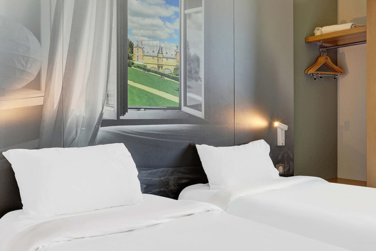 Habitación de hotel con una cama, una ventana que ofrece vistas a un edificio, un perchero de madera y una lámpara fijada a la pared.