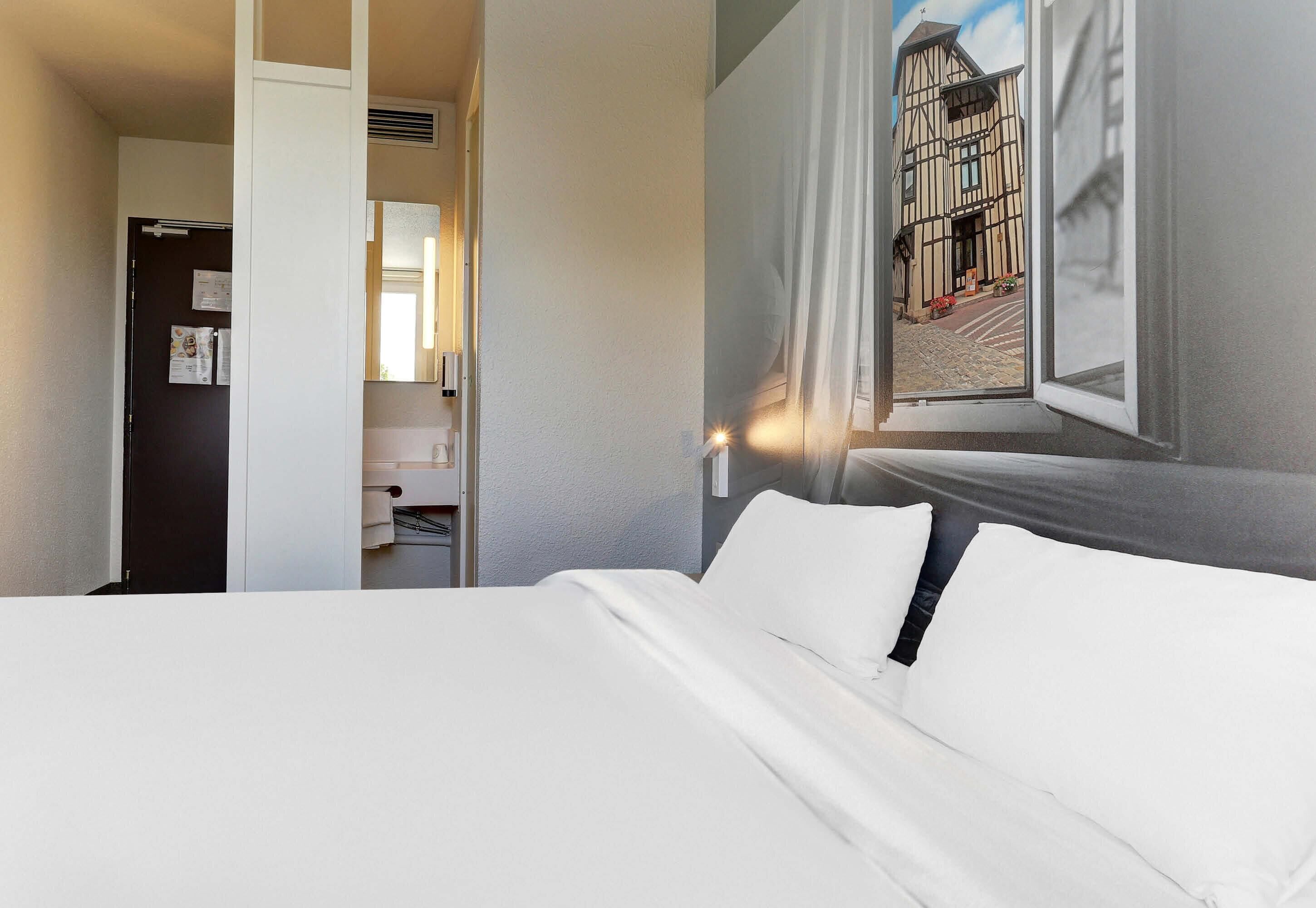 Habitación de hotel con una cama, una ventana con vistas a un edificio, una puerta y un espejo.