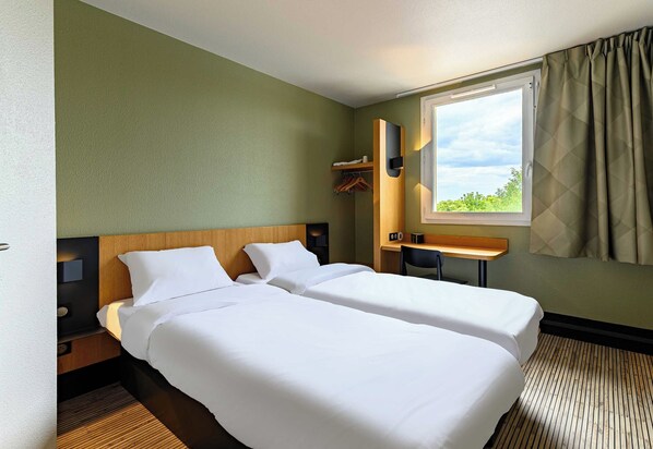 Habitación de hotel con dos camas individuales, un escritorio, una silla, un ventanal con cortinas y un pequeño estante con una toalla y perchas.