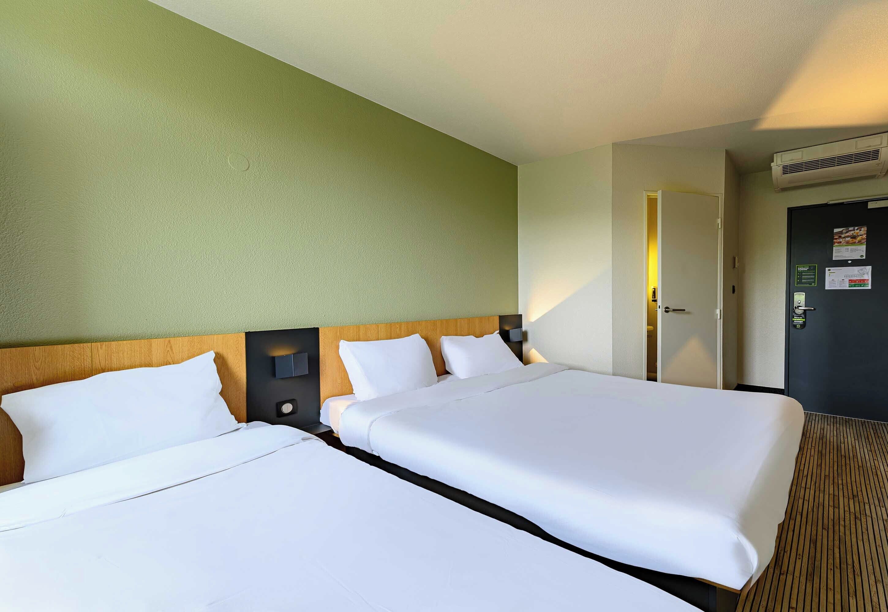 A hotel room with two beds, a wooden headboard, a green wall, and a door with a light switch.