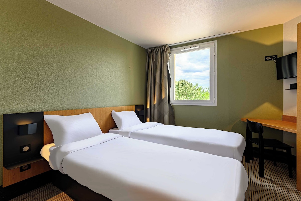 A hotel room with two single beds, a desk, a chair, a TV, and a window with a view of trees.